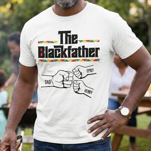 May include: White t-shirt with the text "The Blackfather" in black and red, with a green, yellow, and red design. Below the text are three fists with the names "Dad", "Emily", and "Henry". The shirt is being worn outdoors.