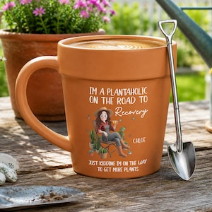 May include: A terracotta-colored ceramic mug shaped like a flower pot, featuring a graphic of a person sitting in a chair surrounded by plants. The text reads: I'M A PLANTAHOLIC ON THE ROAD TO Recovery CHLOE JUST KIDDING I'M ON THE WAY TO GET MORE PLANTS.