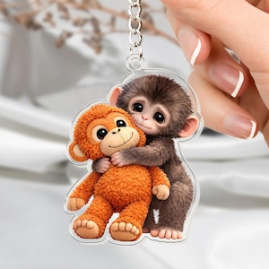 May include: A clear acrylic keychain featuring two cartoon monkeys. One monkey is brown and hugging an orange monkey. The keychain has a silver chain and is held by a hand with manicured nails. The monkeys have large eyes and are smiling.