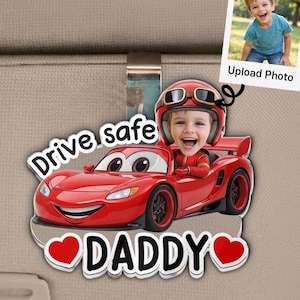 May include: A red car-shaped accessory with the text "Drive safe DADDY" and a photo of a child in a racing helmet. The accessory is designed to be attached to a car's interior. A small photo with the text "Upload Photo" is also included.