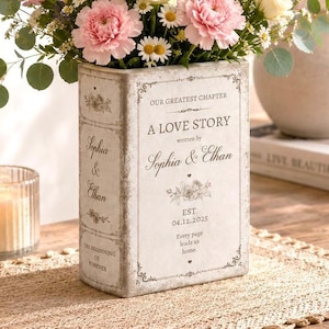 May include: A rectangular ceramic vase shaped like a book, featuring the text "OUR GREATEST CHAPTER A LOVE STORY written by Sophia & Ethan EST. 04.12.2025". The vase is filled with a bouquet of pink roses, white daisies, and purple lavender.