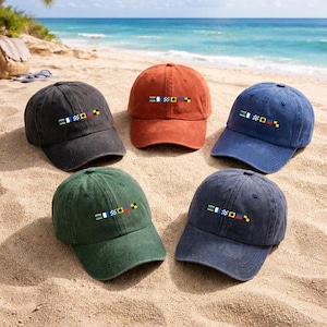 May include: Five baseball caps in different colours: black, rust, blue, green, and navy. Each cap has a small embroidered design with horizontal stripes and squares. The caps are displayed on a sandy beach with the sea in the background.