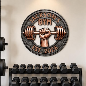 May include: A circular wall sign featuring a copper-coloured fist gripping a barbell, surrounded by a dark textured border. The text reads JACKSON'S GYM and EST. 2026 in copper lettering. The sign is mounted on a wall above a rack of black dumbbells.
