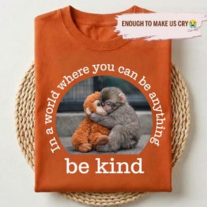 May include: Orange t-shirt with a circular graphic featuring two hugging monkeys and the words "In a world where you can be anything, be kind." The text "ENOUGH TO MAKE US CRY" is above the graphic. The shirt is folded on a woven mat.