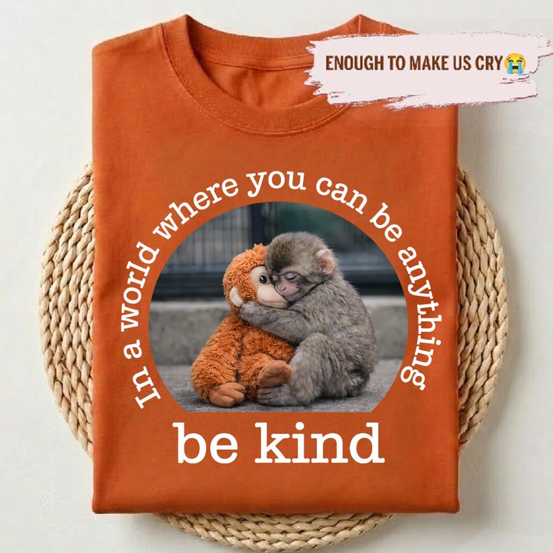 May include: Burnt orange t-shirt featuring a graphic of two hugging monkeys, one plush and one real. The text "In a world where you can be anything, be kind" is printed in a circular design. The phrase "ENOUGH TO MAKE US CRY" is printed at the top.