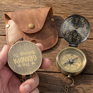 May include: A gold-coloured compass, open to show the directional dial. The compass is engraved with the words "This Belongs To GRANDPA". A brown leather pouch and chain are also visible.