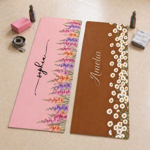 May include: Two rectangular yoga mats. The left mat is pink with a vertical floral pattern of purple, orange, and pink flowers and the name Sophia in black script. The right mat is brown with a vertical pattern of white daisies and the name Amelia.