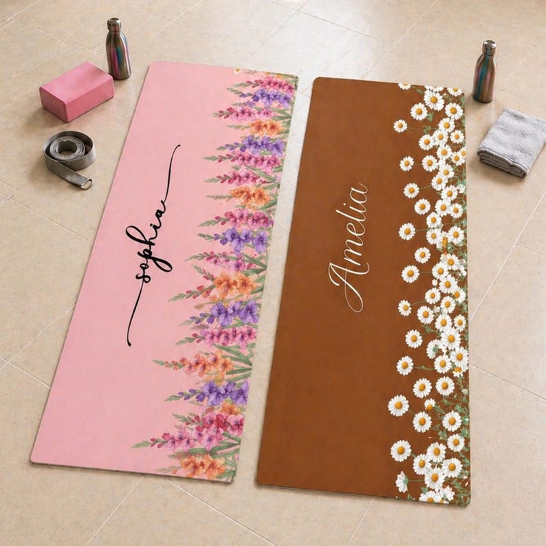 May include: Two rectangular yoga mats. The left mat is pink with a vertical floral pattern of purple, orange, and pink flowers and the name Sophia in black script. The right mat is brown with a vertical pattern of white daisies and the name Amelia.