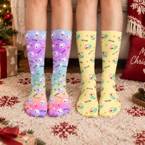May include: Two pairs of crew socks. The left pair features a purple-to-orange gradient background with repeating cartoon cloud faces, red stars, and the name Sophia. The right pair has a yellow background with rainbow cloud faces and the name Olivia.