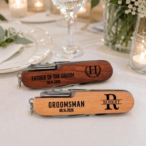 May include: Two multi-tool pocket knives with wooden handles. The top knife is dark wood, engraved with 'FATHER OF THE GROOM', '06.14.2026', and an 'H' in a laurel wreath. The bottom knife is light wood, engraved with 'GROOMSMAN', '06.14.2026', and 'ROBERT'.