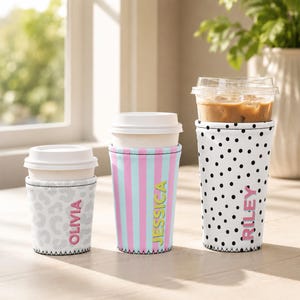 May include: Three beverage cup sleeves with white lids. The first is gray leopard print with the name OLIVIA in pink. The second has vertical pink and blue stripes with the name JESSICA in yellow. The third is white with black polka dots and the name RILEY in pink.