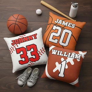 May include: A collection of sports-themed cushions, including basketball, baseball, and American football designs. The cushions feature names and numbers, with a basketball cushion displaying "JAMES 20". A baseball bat, baseball, and trainers are also present.
