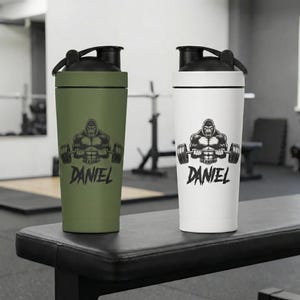 May include: Two shaker bottles, one olive green and one white, each featuring a black lid and a graphic of a muscular gorilla lifting a barbell. Below the gorilla, the name DANIEL is printed in a bold, stylized black font on both bottles.
