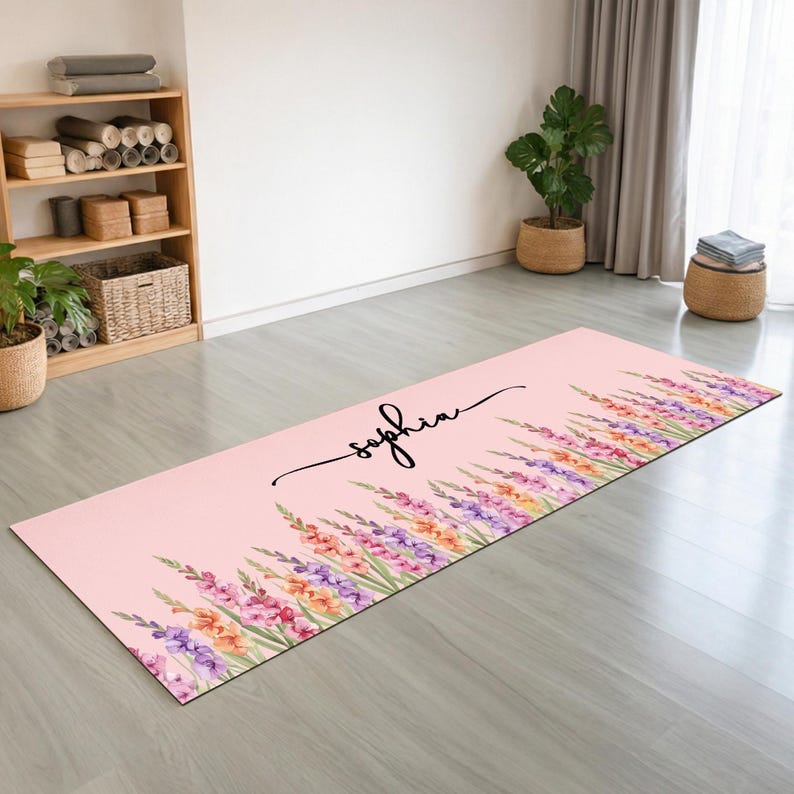 May include: A rectangular yoga mat with a light pink background featuring a watercolor print of colorful gladiolus flowers along the bottom edge and the name Sophia written in black cursive script in the upper center of the mat surface.