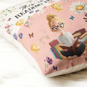 May include: A decorative pillow with a pink and white floral design. The pillow features an illustration of a person reading books, wearing glasses. The text "This is READING PLACE" is printed on the pillow.