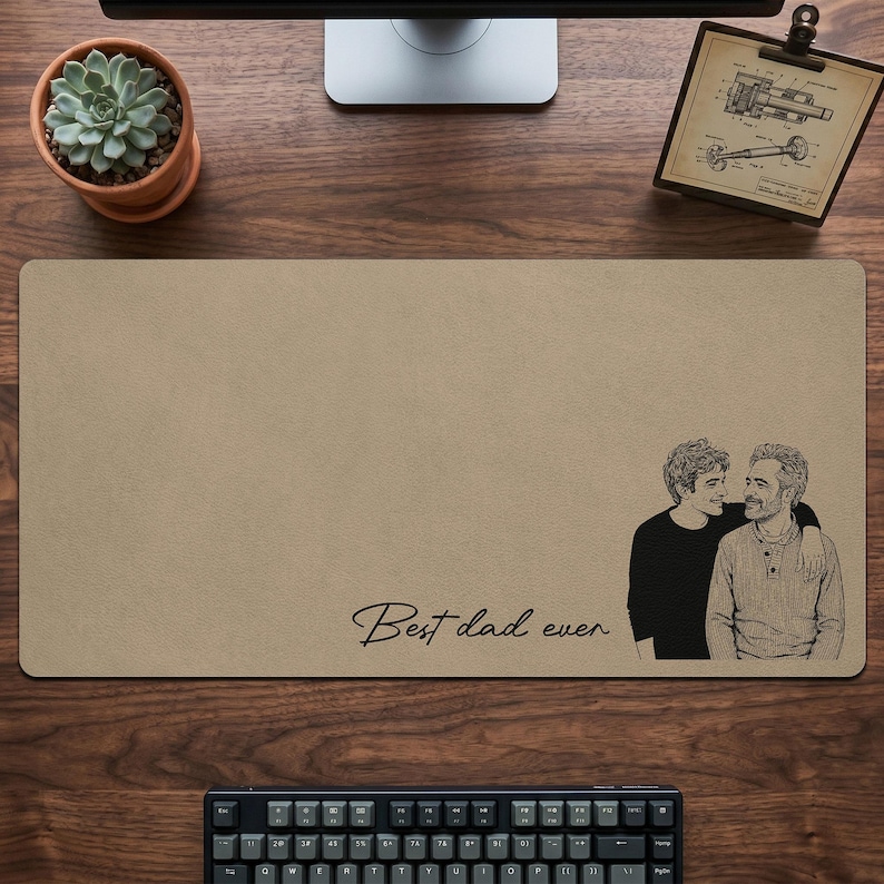 May include: Tan rectangular desk mat with a black and white illustration of two people embracing. The words "Best dad ever" are written in cursive. A succulent plant in a terracotta pot and a keyboard are also visible.