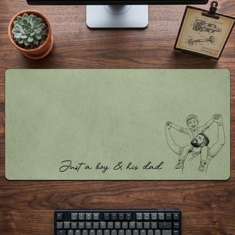 May include: Sage green rectangular desk mat with a black and white illustration of a father and son. The text "Just a boy & his dad" is written in cursive. A keyboard, succulent, and monitor are visible.