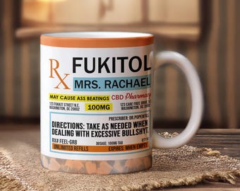 May include: White ceramic mug with an orange rim and a handle. The mug features the text "FUKITOL MRS. RACHAEL" and other text in black and yellow, including "MAY CAUSE ASS BEATINGS" and "DIRECTIONS: TAKE AS NEEDED WHEN DEALING WITH EXCESSIVE BULLSHYT."