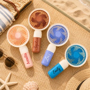 May include: Four portable handheld fans with circular heads and cylindrical handles. Each fan features a unique colour scheme: peach, brown, lavender, and blue. The handles are personalised with the names Amelia, Sophia, Lois, and Emily.