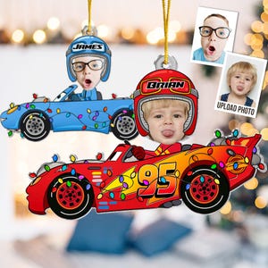May include: Two custom hanging ornaments featuring cartoon race cars decorated with colorful Christmas lights. One blue car displays the name JAMES, and one red car displays the number 95 and the name BRIAN, each with a child's face in the driver seat.