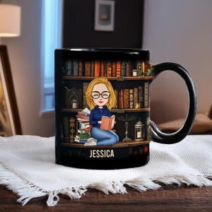 May include: Black ceramic mug featuring a cartoon illustration of a woman reading in front of a bookshelf. The woman has blonde hair and glasses, wearing a blue shirt and jeans. The name "JESSICA" is printed on the mug.