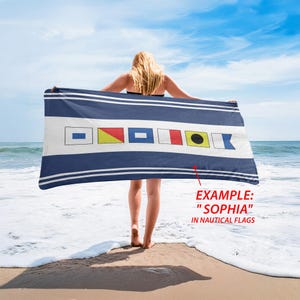 May include: A blue and white nautical beach towel featuring signal flags and horizontal stripes. The towel is displayed on a beach with the sea and sky visible. The text "EXAMPLE: 'SOPHIA' IN NAUTICAL FLAGS" is shown.