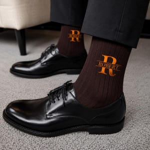 May include: A pair of dark brown ribbed socks featuring an embroidered orange monogram with a large letter R and the name ROBERT centered horizontally across the letter. The socks are worn with black leather dress shoes and black trousers.