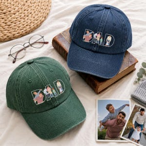May include: Two baseball caps, one dark green and one navy blue, are displayed. Each cap features the word "DAD" with a family illustration. The caps are on a white surface with a book, glasses, and photos.