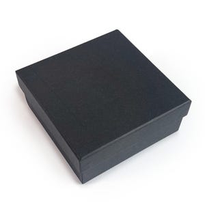 May include: A black, rectangular gift box with a textured surface. The box is closed and has a lid that fits snugly over the base. The box is set against a white background.