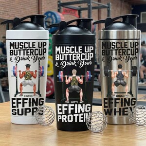 Puede incluir: Tres botellas de coctelera de acero inoxidable con tapas negras. Cada botella presenta un gr&aacute;fico de levantador de pesas y el texto "Muscle Up Buttercup & Drink Your". Una botella es blanca, una negra y otra plateada.