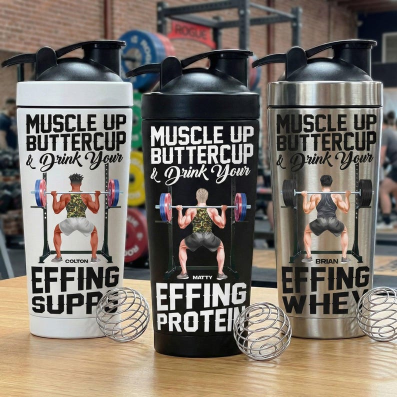 Puede incluir: Tres botellas de coctelera de acero inoxidable con tapas negras. Cada botella presenta un gr&aacute;fico de levantador de pesas y el texto "Muscle Up Buttercup & Drink Your". Una botella es blanca, una negra y otra plateada.