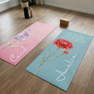 May include: Two yoga mats with floral designs and personalized names. One mat is pink with a lavender illustration and the name "Carol". The other is blue with a red carnation and the name "Amelia". Both mats have the text "May I Be Safe, May I Be Happy, May I Be Healthy, May I Live With Ease."