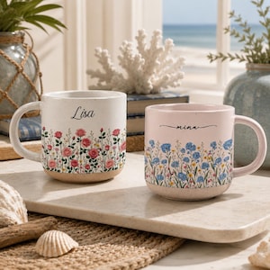 May include: Two ceramic mugs with speckled finishes. The left mug is white with pink roses and the name Lisa. The right mug is light pink with blue wildflowers and the name mina. Both feature a handle and a natural, unglazed base texture.