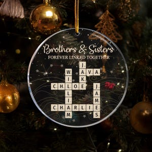 May include: A clear, circular Christmas decoration inscribed with "Brothers & Sisters FOREVER LINKED TOGETHER." The ornament has a crossword design with names in Scrabble tiles. It hangs from a gold ribbon against a Christmas tree background.