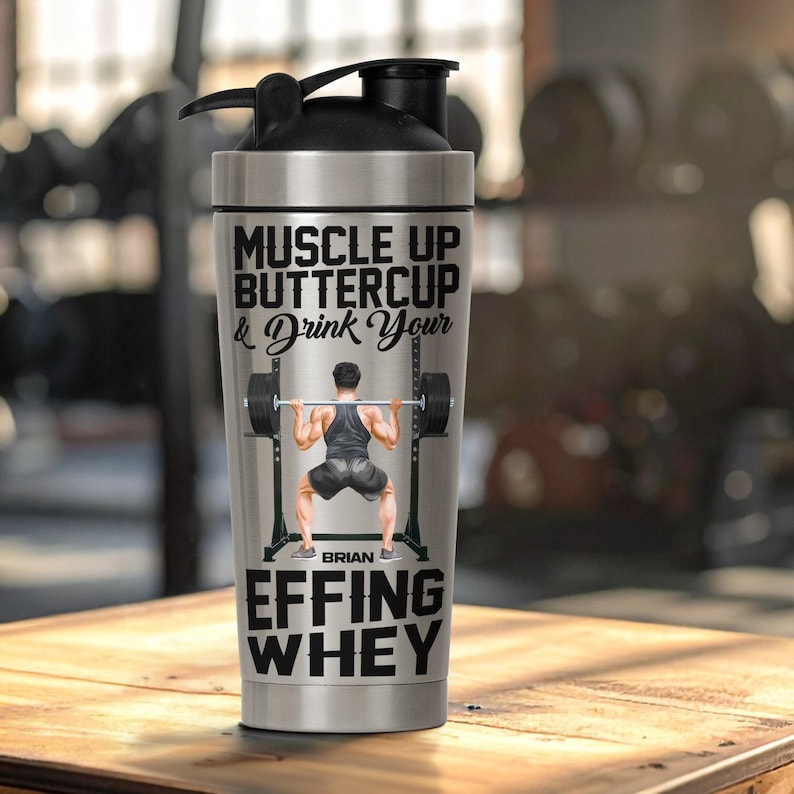 Puede incluir: Una botella mezcladora de acero inoxidable con tapa negra. La botella presenta el texto "MUSCLE UP BUTTERCUP & Drink Your" y "EFFING WHEY". Un gr&aacute;fico de un levantador de pesas tambi&eacute;n est&aacute; en la botella. El nombre "BRIAN" est&aacute; impreso debajo.