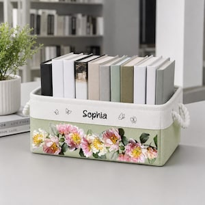May include: A rectangular fabric storage bin with white rope handles, featuring a sage green base with a floral print of pink and yellow flowers. The white upper band displays the name Sophia in black script, flanked by small black butterfly outlines.