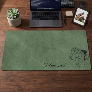 May include: A rectangular, sage green desk mat with a textured surface. The mat features a black line drawing of a person holding a child and the handwritten text "I love you!". A laptop, succulent, and framed photo are also on the wooden desk.