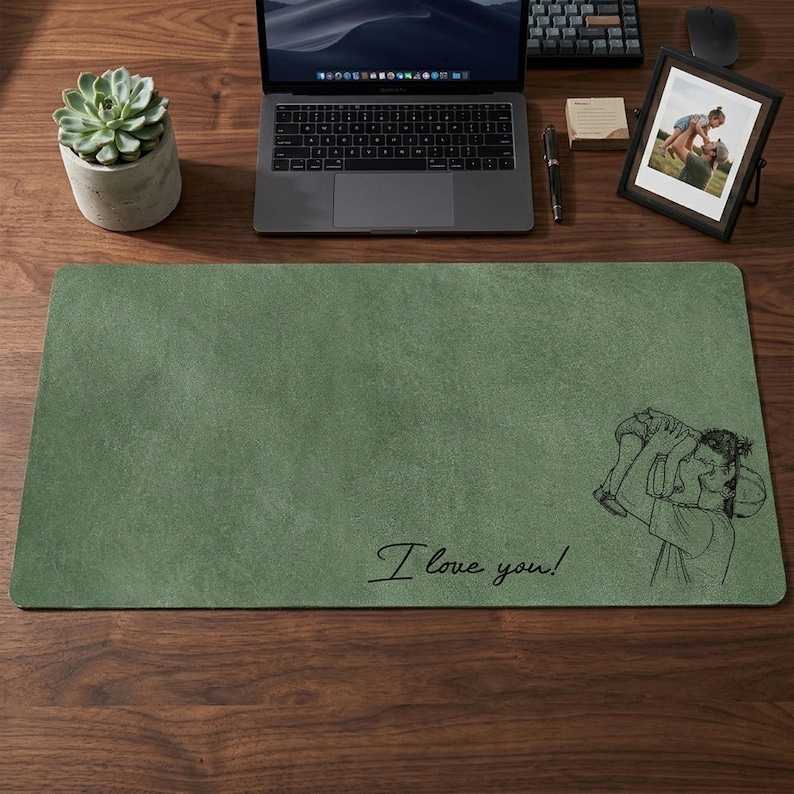 May include: A rectangular, sage green desk mat with a textured surface. The mat features a black line drawing of a person holding a child and the handwritten text "I love you!". A laptop, succulent, and framed photo are also on the wooden desk.