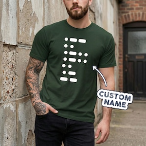 May include: Dark green t-shirt with a white Morse code design. The design is a series of dots and dashes forming the letter "F". The shirt is a crew neck style and is made of a soft material. The words "CUSTOM NAME" are in a white box.