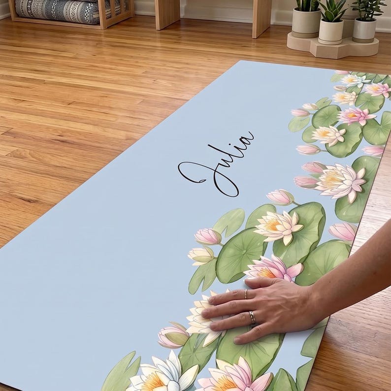 May include: A light blue rectangular yoga mat featuring a printed pattern of green lily pads and white and pink water lilies along the right side. The name Julia is printed in black cursive script in the center of the mat surface.