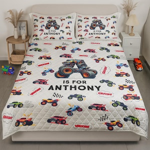May include: A white quilt and two pillowcases featuring a monster truck theme. The quilt has a repeating pattern of colorful monster trucks, checkered flags, and speech bubbles with the words "CRASH" and "VROOM". The text "IS FOR ANTHONY" is prominently displayed.