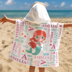May include: A white hooded beach towel featuring a mermaid illustration with red hair and a teal tail. The towel is personalized with the name "Amelia" in pink and teal, along with starfish and seashell graphics.