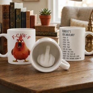 May include: Three white ceramic mugs. One mug features a red chicken with the text F-CAW-F. Another mug displays the text ROBERT'S VOCABULARY followed by a list of profanities. The third mug is tilted to reveal a white hand sculpture showing a middle finger.