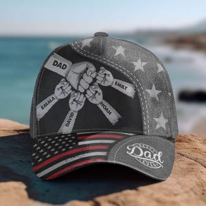 May include: A gray baseball cap with a design featuring fists and the names "Emma", "David", "Noah", and "Emily". The cap also has the text "DAD" and "Best Dad Ever" and an American flag design on the brim.