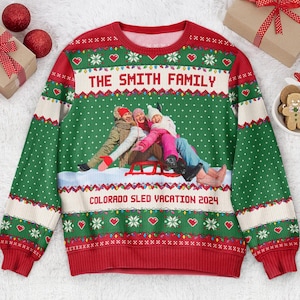 May include: A green and red long-sleeve crewneck jumper featuring a repeating pattern of white dots, hearts, and snowflakes. The centre displays a photograph of three people on a red sled with the text THE SMITH FAMILY and COLORADO SLED VACATION 2024.
