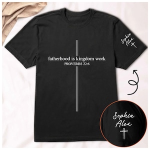 May include: Black t-shirt with the text "fatherhood is kingdom work PROVERBS 22:6" in white. A white vertical line and cross design is centered on the shirt. The sleeve has the name "Sophia Alex" and a cross.