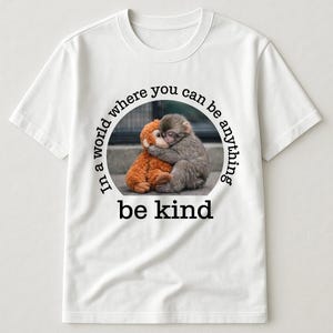 May include: White t-shirt featuring a circular graphic with a brown stuffed monkey hugging a gray monkey. The text "In a world where you can be anything, be kind" is printed around the image in black.