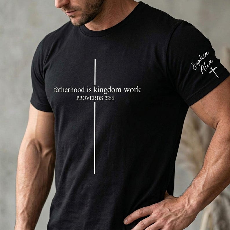 May include: Black crew neck t-shirt with white text and a vertical line forming a cross. The text reads "fatherhood is kingdom work" and "PROVERBS 22:6". The sleeve has the text "Sophia Alex" and a cross.
