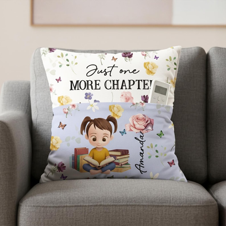 May include: A cushion with a pocket, featuring a cartoon girl reading books. The pillow has a floral design with butterflies and the text "Just one MORE CHAPTER!". A remote control is in the pocket.
