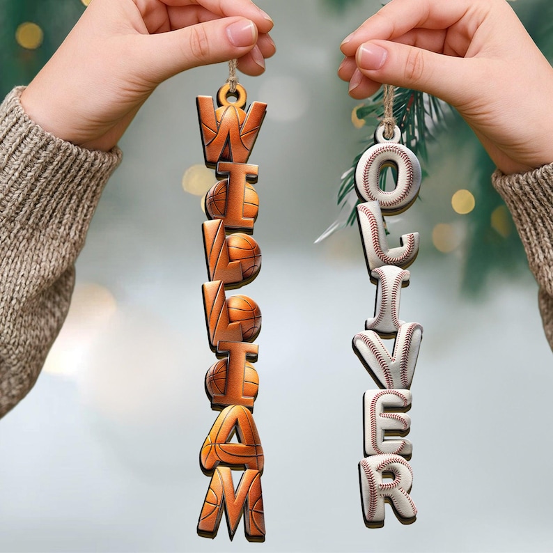 May include: Two wooden ornaments are held up by hands. One ornament spells "WILLIAM" with basketball-themed letters. The other spells "OLIVER" with baseball-themed letters. Both are suspended by twine.